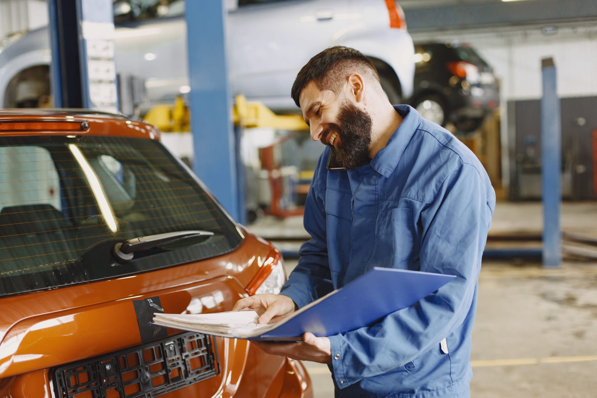 Mechanic in blue coveralls reviewing a work order beside a car in a service garage