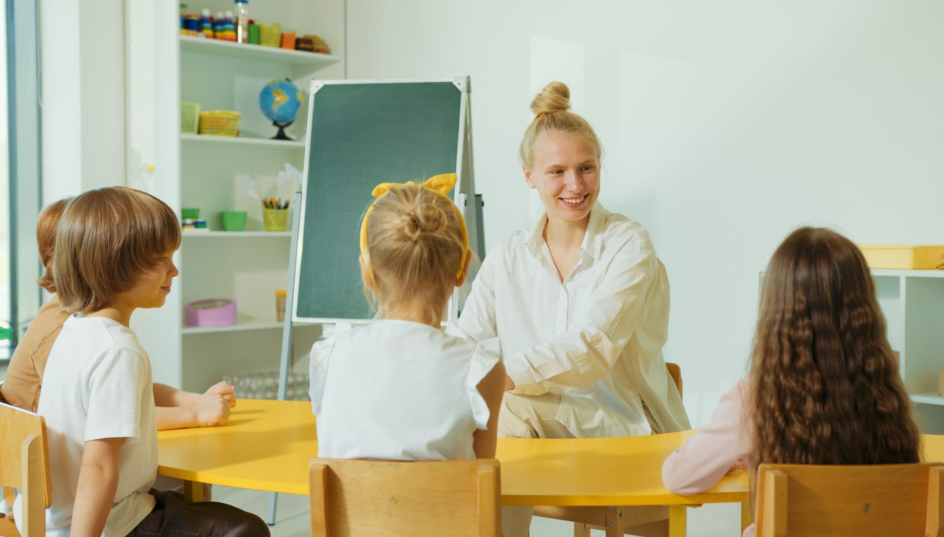 Preschool teacher at a chalkboard engaging young children seated at a classroom table